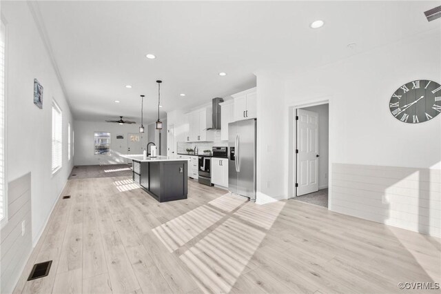 kitchen featuring stainless steel appliances, wall chimney range hood, pendant lighting, a center island with sink, and white cabinets