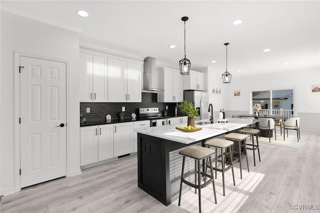 kitchen with appliances with stainless steel finishes, wall chimney exhaust hood, pendant lighting, white cabinetry, and an island with sink