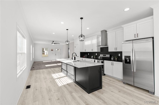 kitchen featuring a center island with sink, wall chimney range hood, appliances with stainless steel finishes, decorative light fixtures, and white cabinetry