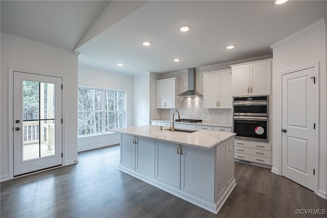 kitchen featuring wall chimney exhaust hood, double oven, sink, white cabinets, and an island with sink