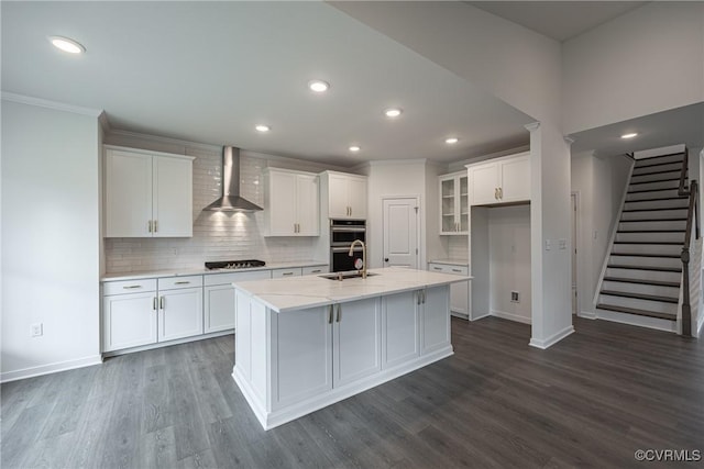 kitchen featuring double oven, white cabinetry, an island with sink, and wall chimney range hood