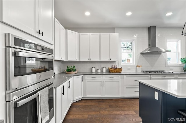 kitchen with decorative backsplash, appliances with stainless steel finishes, dark hardwood / wood-style flooring, wall chimney exhaust hood, and white cabinetry