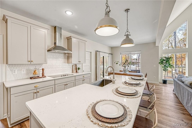 kitchen with sink, wall chimney range hood, light stone counters, an island with sink, and decorative light fixtures