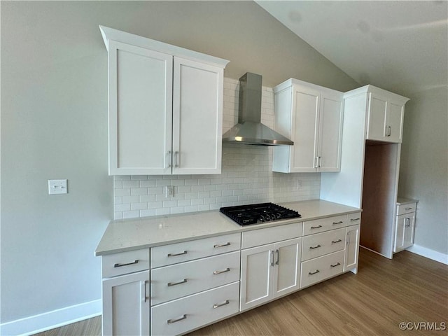 kitchen with wall chimney range hood, tasteful backsplash, baseboards, white cabinetry, and vaulted ceiling