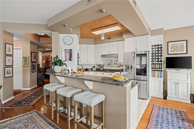 kitchen featuring dark stone counters, a kitchen breakfast bar, stainless steel refrigerator with ice dispenser, hanging light fixtures, and white cabinetry