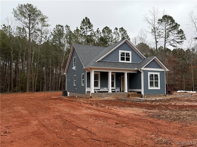 view of front of house featuring a shingled roof, covered porch, a forest view, and central AC unit