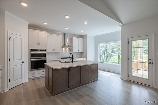 kitchen featuring a kitchen island with sink, white cabinets, wall chimney range hood, sink, and stainless steel double oven