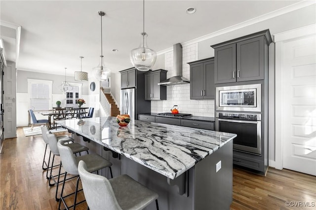 kitchen featuring wall chimney exhaust hood, a kitchen bar, stainless steel appliances, hanging light fixtures, and a large island