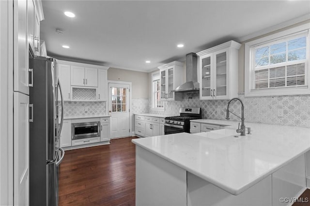 kitchen with kitchen peninsula, stainless steel appliances, wall chimney range hood, dark hardwood / wood-style flooring, and white cabinetry