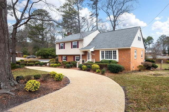 tri-level home with driveway, a front yard, a shingled roof, brick siding, and a chimney