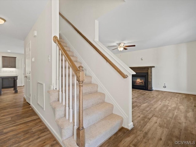 stairs featuring baseboards, visible vents, a glass covered fireplace, ceiling fan, and wood finished floors