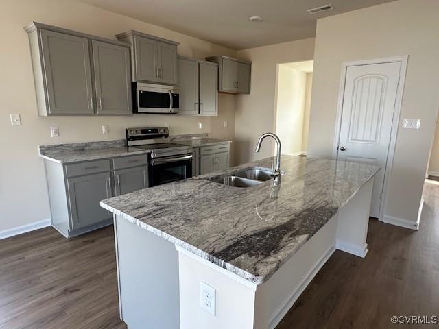 kitchen with appliances with stainless steel finishes, dark hardwood / wood-style floors, sink, light stone countertops, and a center island with sink