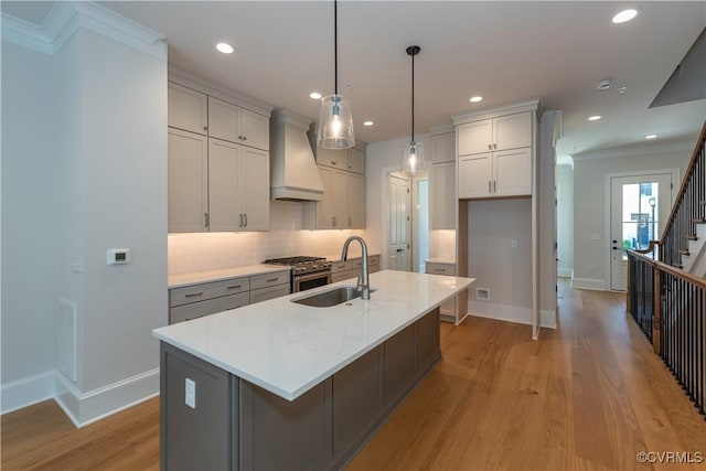 kitchen featuring pendant lighting, custom exhaust hood, a center island with sink, light hardwood / wood-style floors, and light stone counters