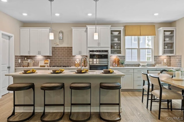 kitchen featuring white cabinets, stainless steel double oven, hanging light fixtures, and an island with sink