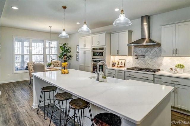 kitchen featuring appliances with stainless steel finishes, a large island with sink, pendant lighting, decorative backsplash, and wall chimney exhaust hood