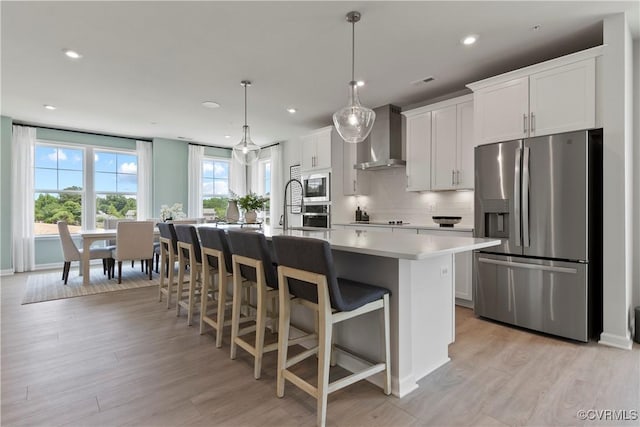 kitchen with a center island with sink, stainless steel appliances, hanging light fixtures, wall chimney exhaust hood, and white cabinetry