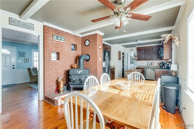 dining space with a wood stove, ceiling fan, light hardwood / wood-style floors, ornamental molding, and beam ceiling