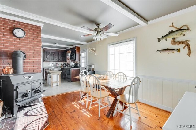 dining space featuring light hardwood / wood-style floors, beamed ceiling, ceiling fan, ornamental molding, and a wood stove