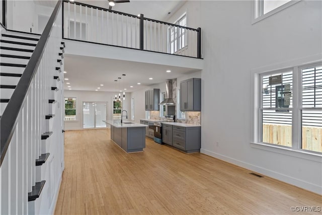 kitchen featuring stainless steel range with electric stovetop, gray cabinets, a high ceiling, a kitchen island with sink, and wall chimney exhaust hood