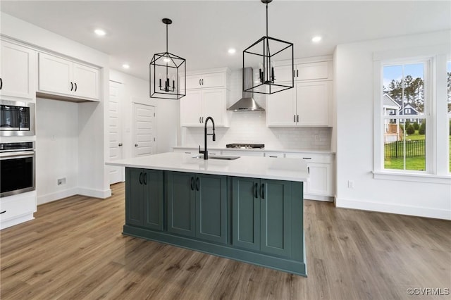 kitchen featuring pendant lighting, white cabinets, wall chimney exhaust hood, stainless steel appliances, and an island with sink