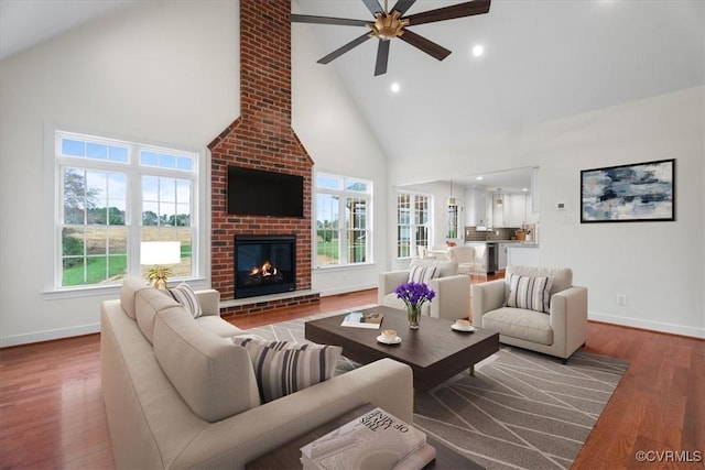 living room featuring high vaulted ceiling, a healthy amount of sunlight, wood-type flooring, and a brick fireplace