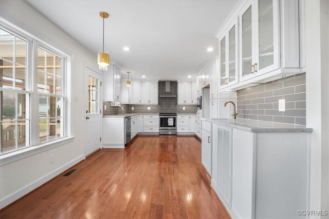 kitchen with pendant lighting, white cabinets, wall chimney exhaust hood, stainless steel appliances, and light stone counters