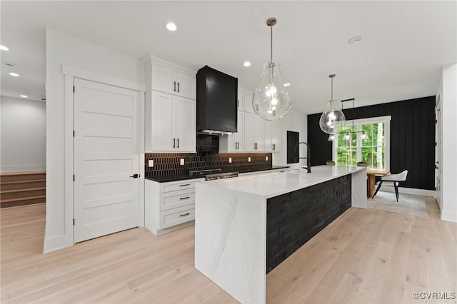 kitchen with light wood finished floors, a large island, wall chimney range hood, white cabinetry, and backsplash