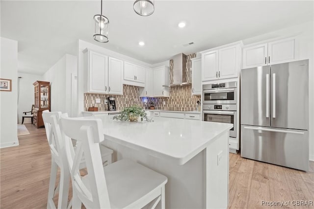 kitchen featuring wall chimney exhaust hood, white cabinetry, stainless steel appliances, light wood-type flooring, and a breakfast bar