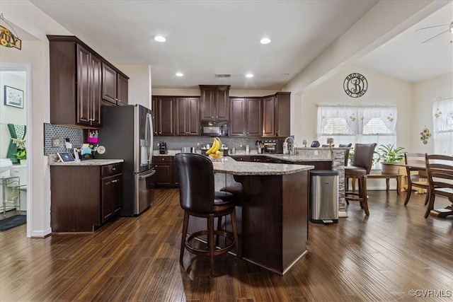 kitchen featuring vaulted ceiling, a breakfast bar area, stainless steel fridge, light stone counters, and dark wood-type flooring