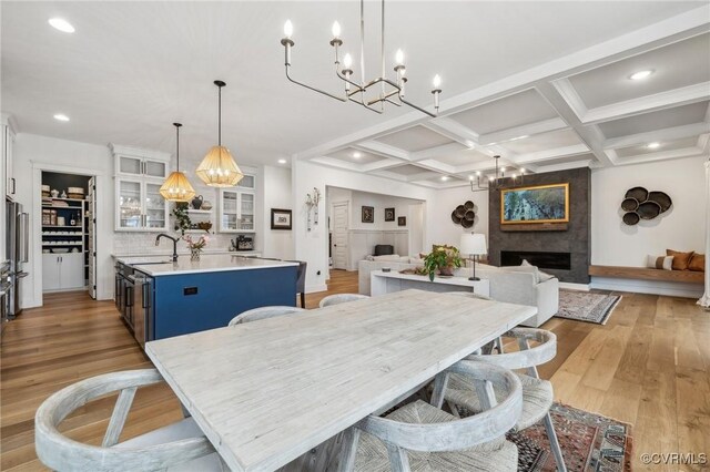 dining space featuring light wood-style floors, a large fireplace, a chandelier, coffered ceiling, and beamed ceiling