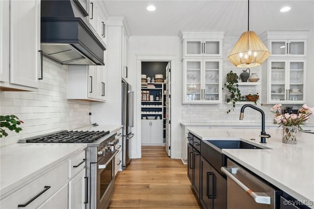 kitchen with custom range hood, appliances with stainless steel finishes, glass insert cabinets, hanging light fixtures, and a sink