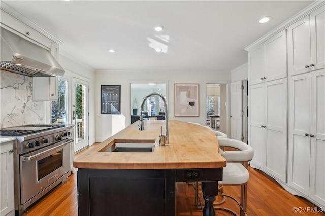 kitchen with wooden counters, extractor fan, an island with sink, decorative backsplash, and high end range