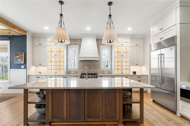 kitchen with built in fridge, white cabinetry, custom range hood, and a kitchen island with sink