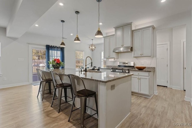 kitchen featuring under cabinet range hood, a sink, stainless steel range oven, light wood finished floors, and an island with sink