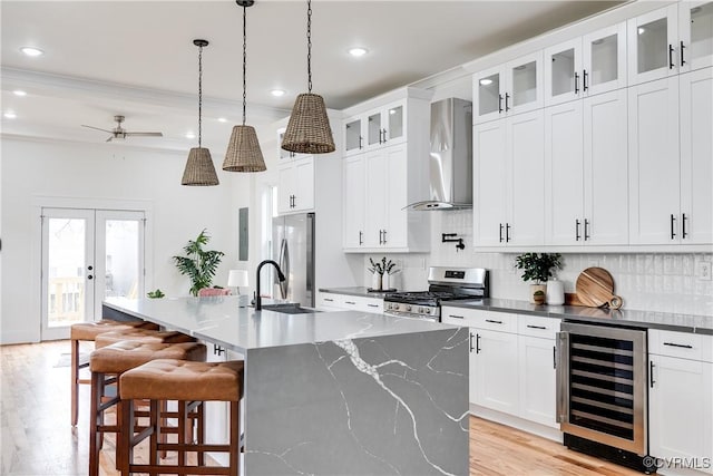 kitchen featuring white cabinetry, appliances with stainless steel finishes, an island with sink, beverage cooler, and wall chimney range hood