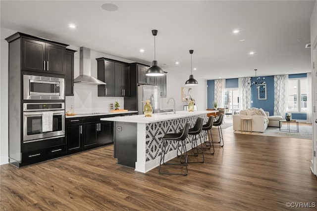 kitchen with wall chimney exhaust hood, a breakfast bar, hanging light fixtures, appliances with stainless steel finishes, and an island with sink