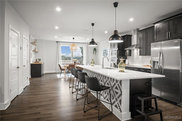 kitchen featuring appliances with stainless steel finishes, hanging light fixtures, a kitchen breakfast bar, a center island with sink, and wall chimney exhaust hood