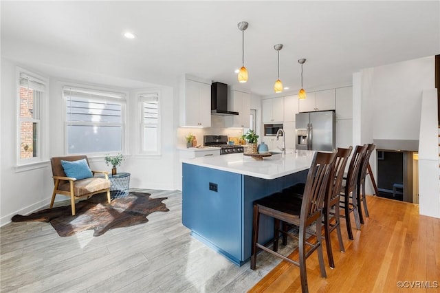 kitchen featuring pendant lighting, appliances with stainless steel finishes, white cabinets, a center island with sink, and wall chimney exhaust hood