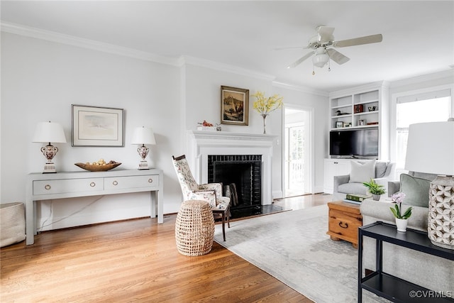 living room featuring built in shelves, light hardwood / wood-style flooring, ornamental molding, ceiling fan, and a fireplace