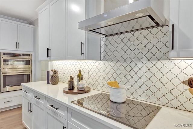 kitchen featuring white cabinetry, double oven, light stone countertops, decorative backsplash, and wall chimney range hood