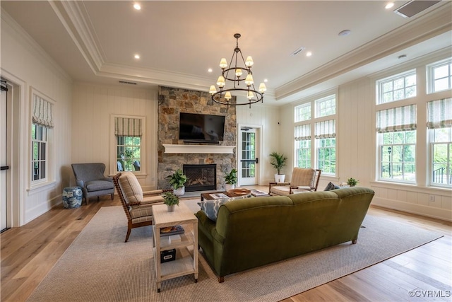 living room with crown molding, light hardwood / wood-style floors, a stone fireplace, a raised ceiling, and a chandelier