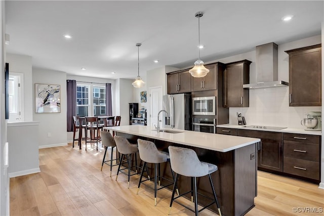 kitchen featuring an island with sink, appliances with stainless steel finishes, wall chimney range hood, and decorative light fixtures