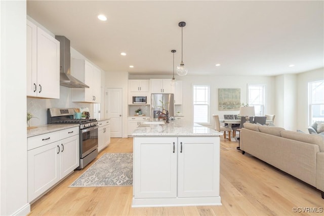 kitchen featuring wall chimney range hood, hanging light fixtures, stainless steel appliances, an island with sink, and white cabinets