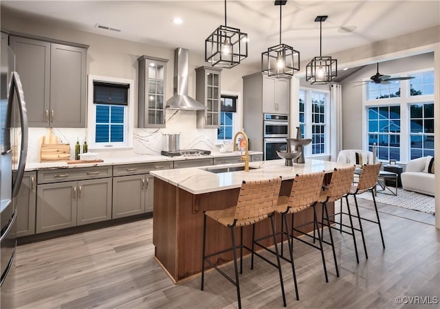 kitchen featuring sink, decorative light fixtures, a center island with sink, gray cabinets, and wall chimney range hood