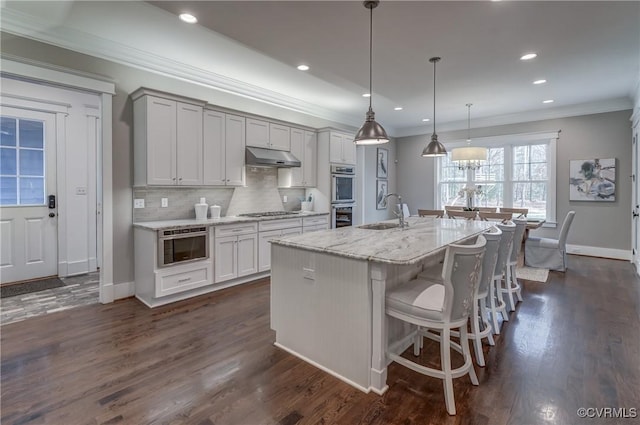 kitchen with sink, hanging light fixtures, appliances with stainless steel finishes, an island with sink, and light stone countertops