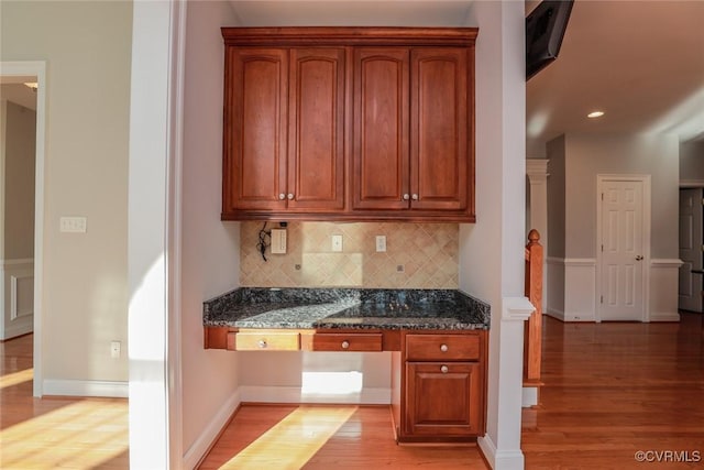 kitchen with backsplash, dark stone countertops, built in desk, and light wood-type flooring