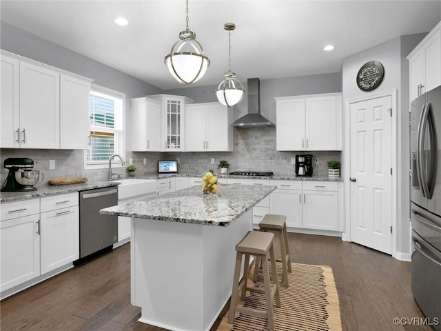 kitchen with appliances with stainless steel finishes, decorative light fixtures, white cabinets, a center island, and wall chimney range hood