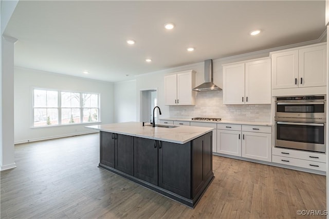 kitchen with sink, white cabinetry, stainless steel appliances, a kitchen island with sink, and wall chimney range hood