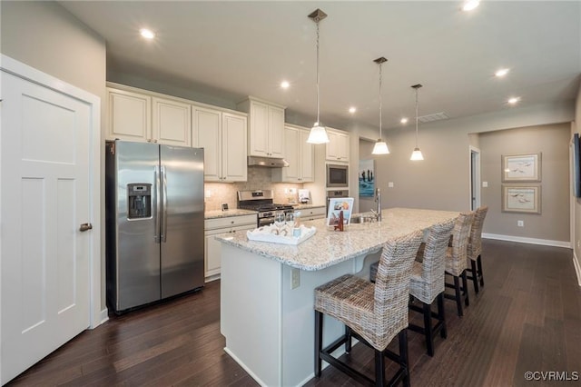 kitchen featuring a kitchen bar, light stone counters, hanging light fixtures, stainless steel appliances, and a kitchen island with sink