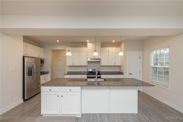 kitchen featuring white cabinets, dark stone counters, hanging light fixtures, stainless steel appliances, and a center island with sink